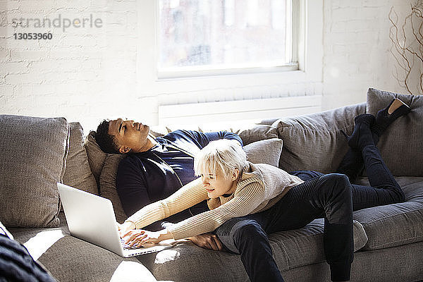 Junge Frau benutzt Laptop  während sie auf einem Mann liegt  der zu Hause auf dem Sofa sitzt