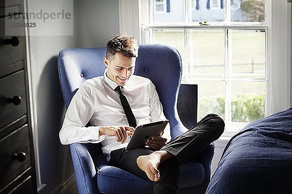 Glücklicher Mann benutzt Tablet-Computer  während er sich zu Hause auf einem Sessel entspannt