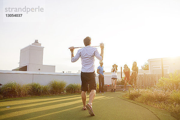 Freunde betrachten einen Mann mit einem Golfschläger  der bei Sonnenuntergang auf der Terrasse des Gebäudes steht
