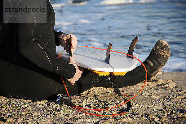 Niedriger Abschnitt eines Surfers  der am Strand sitzend ein Surfbrett-Seil vorbereitet