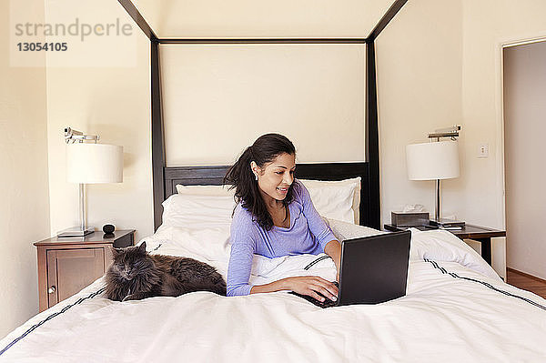 Glückliche Frau liegt neben der Katze und benutzt den Laptop zu Hause auf dem Bett