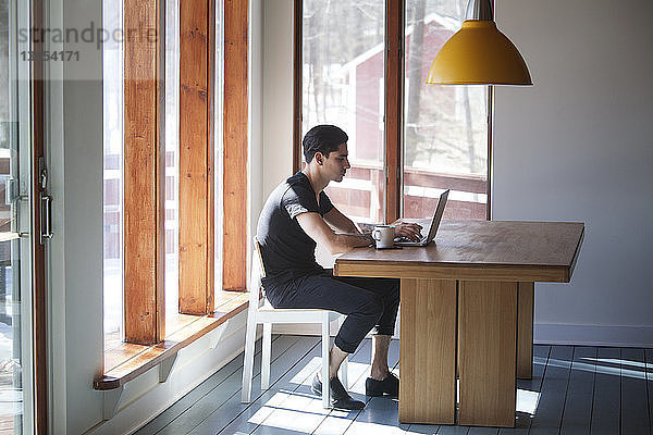 Seitenansicht eines Mannes  der einen Laptop-Computer benutzt  während er zu Hause auf einem Stuhl am Tisch sitzt