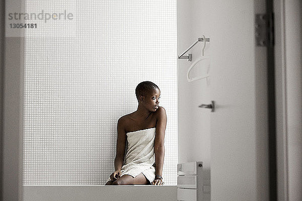 In ein Handtuch gewickelte Frau schaut weg  während sie zu Hause auf der Badewanne sitzt