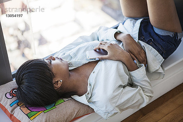 Hochwinkelansicht einer Frau  die schläft  während sie zu Hause ein Smartphone am Fenster hält