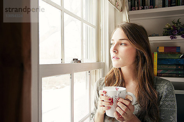 Nachdenkliche Frau hält Kaffeetasse  während sie zu Hause durchs Fenster schaut