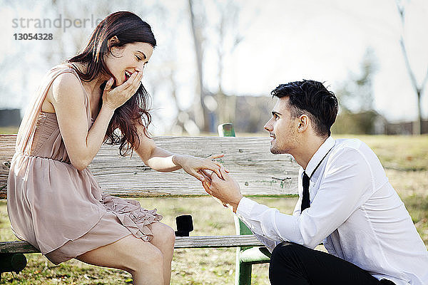 Glückliche Frau sitzt auf Bank  während ein Mann im Park einen Heiratsantrag macht