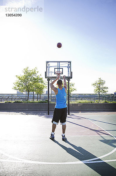 Rückansicht eines Mannes  der an einem sonnigen Tag Basketball im Reifen gegen den Himmel wirft