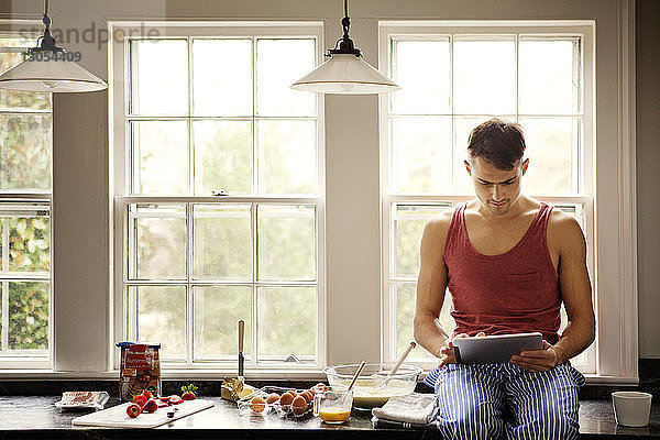 Mann benutzt Tablet-Computer für die Rezeptsuche  während er zu Hause auf der Küchentheke sitzt