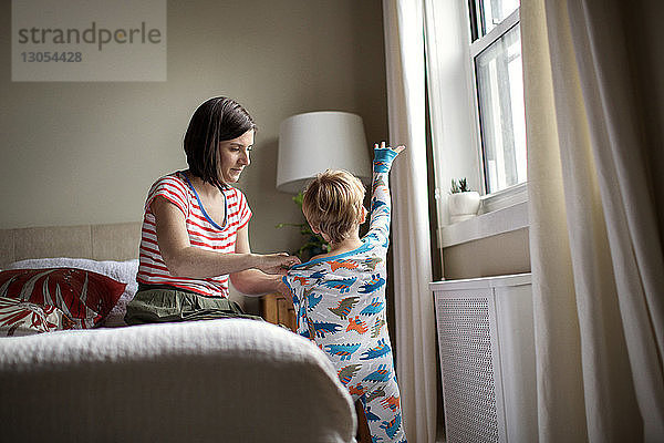 Mutter zieht Sohn an  während sie im Zimmer auf dem Bett sitzt