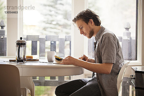 Mann liest Zeitschrift  während er zu Hause auf einem Stuhl am Frühstückstisch sitzt