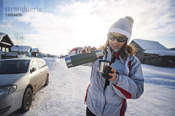 Glückliche Frau gießt Getränk aus Isolierkanne aus  während sie auf schneebedecktem Feld steht