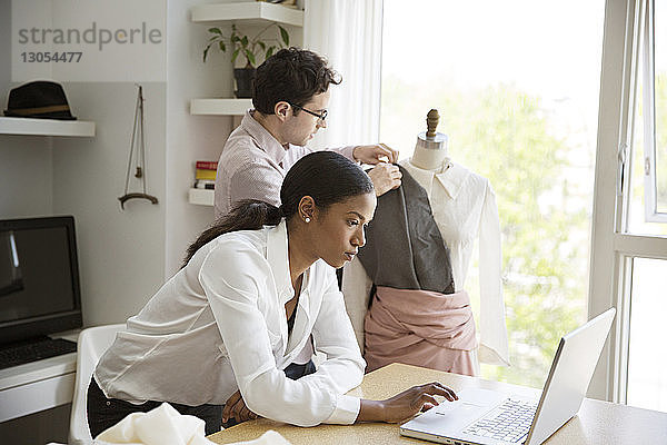 Frau arbeitet am Laptop  während ein Kollege im Atelier ein Modell mit einem Kleid bekleidet