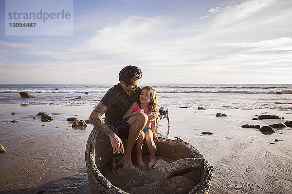 Vater und Tochter sitzen bei Sonnenuntergang auf einem Betonrohr am Strand