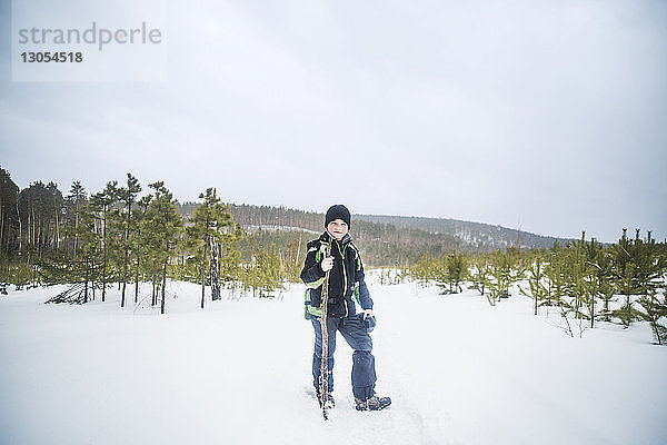 Porträt eines Jungen  der einen Stock hält  während er auf einem schneebedeckten Feld vor klarem Himmel steht