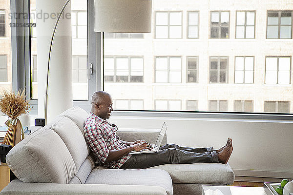 Seitenansicht eines Mannes  der einen Laptop-Computer benutzt  während er zu Hause auf dem Sofa sitzt
