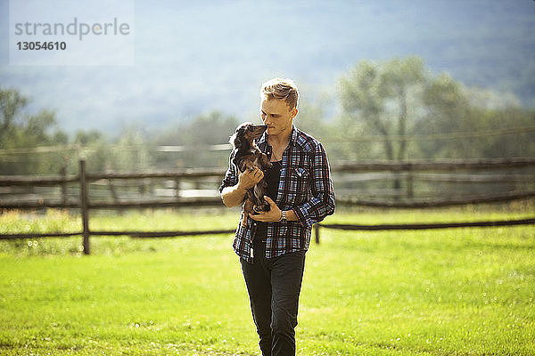 Mann trägt Hund beim Spaziergang auf Grasfeld in Bauernhof