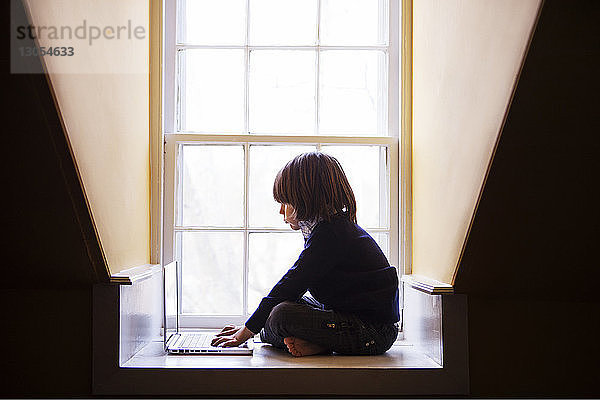 Junge benutzt Laptop-Computer  während er zu Hause am Fenster sitzt