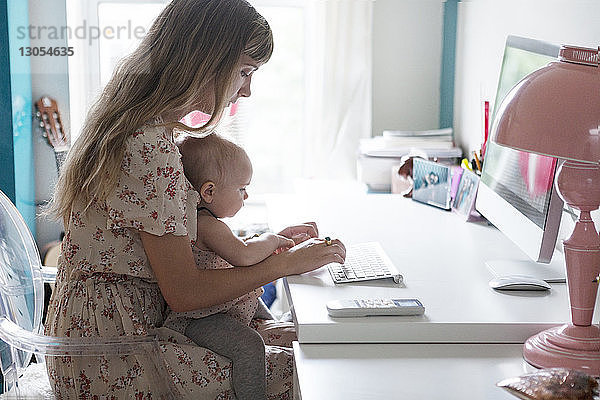 Mutter  die ihr Baby hält  während sie zu Hause einen Desktop-Computer benutzt