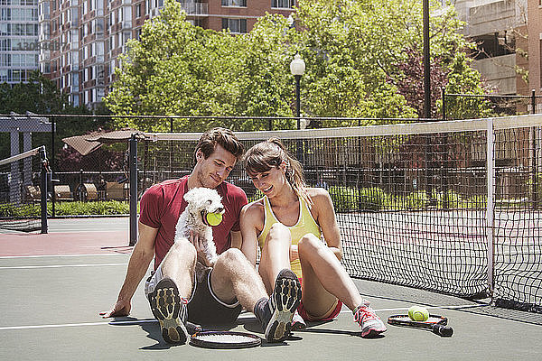 Glückliches Paar sitzt mit Hund auf dem Tennisplatz