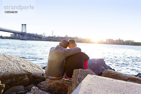 Rückansicht eines Mannes  der bei Sonnenuntergang mit seinem Freund auf Felsen am East River sitzt
