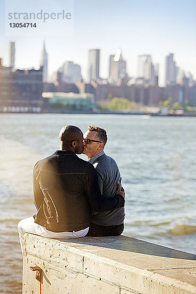 Rückansicht von schwulen Männern  die sich küssen  während sie auf einer Stützmauer am Fluss sitzen