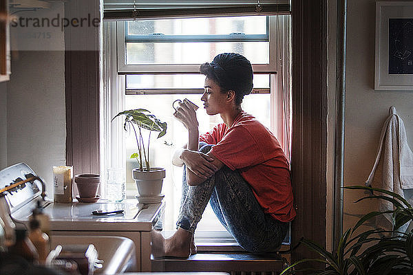 Seitenansicht einer Frau  die trinkt  während sie zu Hause auf dem Fensterbrett sitzt