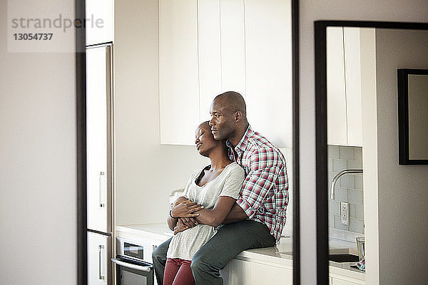 Mann umarmt Frau  während er zu Hause auf dem Küchentisch sitzt