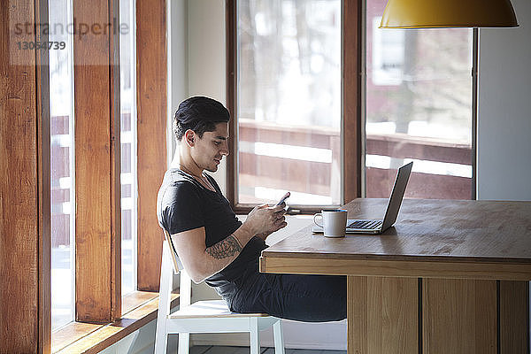 Mann benutzt ein Smartphone  während er zu Hause auf einem Stuhl am Tisch sitzt