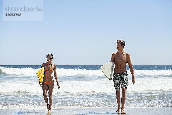 Glückliches Paar trägt Surfbrett beim Strandspaziergang vor klarem Himmel