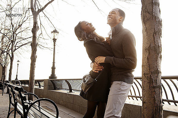 Fröhliches Paar umarmt sich an einem sonnigen Tag am Geländer gegen den Himmel