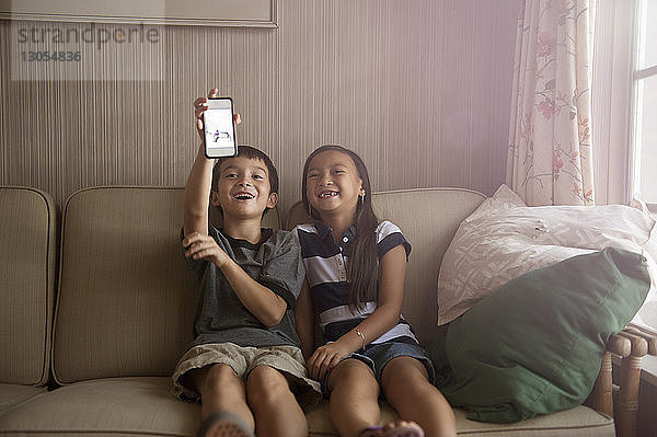 Glückliche Geschwister halten Handy in der Hand  während sie auf dem Sofa sitzen