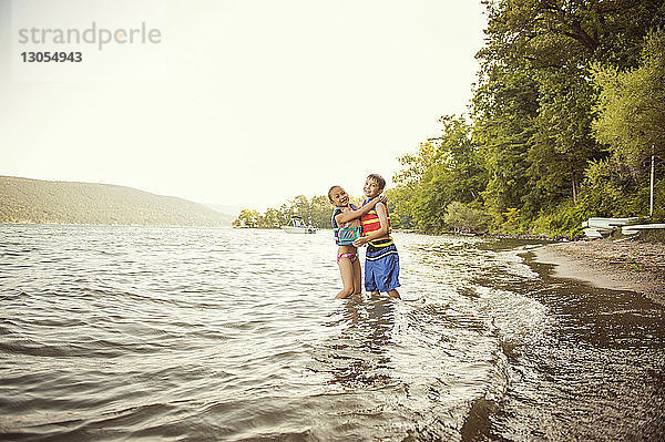 Fröhliche Geschwister umarmen sich  während sie Schwimmwesten im See vor klarem Himmel tragen