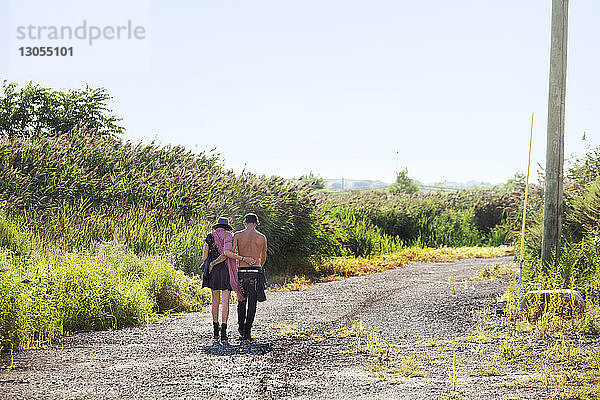 Rückansicht eines Paares mit umgebundenen Armen  das auf einer Schotterstraße durch ein Feld läuft