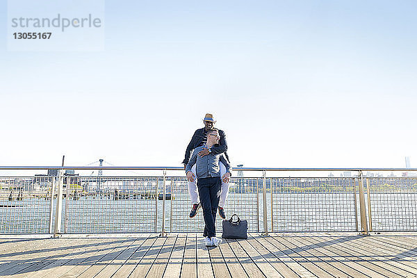 Glücklicher Mann umarmt Freund auf Pier am Fluss bei klarem Himmel