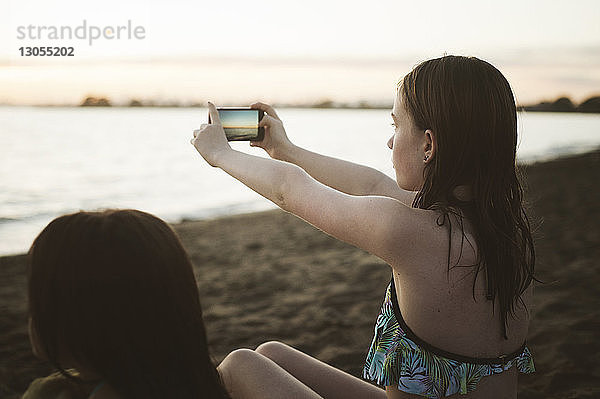 Mädchen fotografiert mit dem Handy  während sie mit ihrer Schwester am Strand sitzt