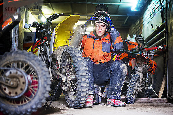 Porträt eines männlichen Motorradfahrers  der einen Helm hält und in einer Garage sitzt