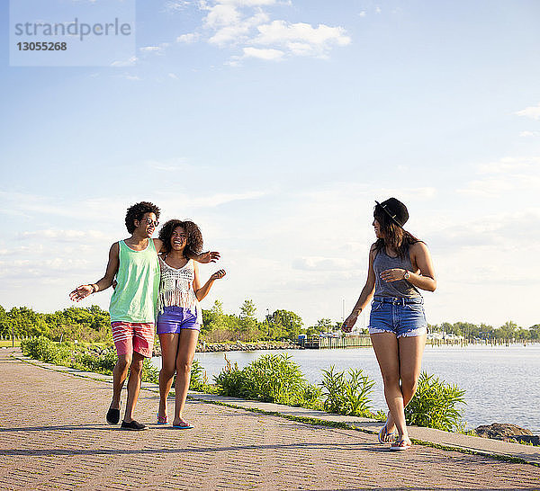 Glückliche Freunde  die an einem sonnigen Tag auf einem Fußweg am Fluss gegen den Himmel laufen