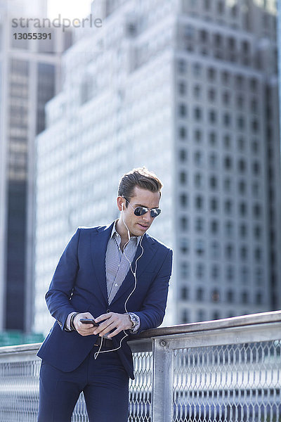 Geschäftsmann hört Musik am Telefon  während er sich in der Stadt an ein Geländer lehnt