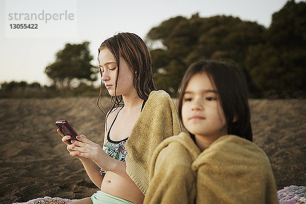 Mädchen benutzt Mobiltelefon  während sie mit ihrer Schwester am Strand sitzt