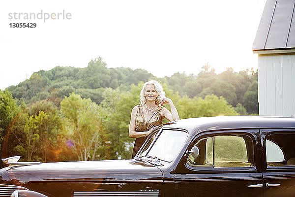 Porträt einer glücklichen älteren Frau  die an einem Oldtimer vor Bäumen steht