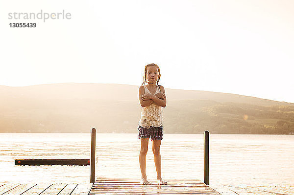 Porträt eines Mädchens mit verschränkten Armen  das bei Sonnenuntergang auf einem Steg über dem See vor klarem Himmel steht