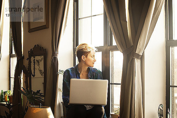 Frau mit Laptop-Computer sitzt zu Hause auf der Fensterbank