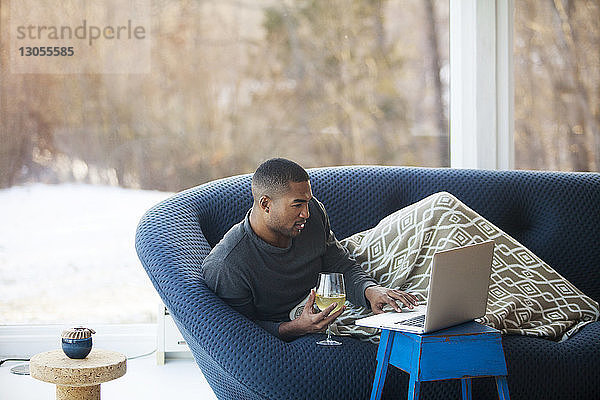 Mann mit Weinglas benutzt Laptop-Computer  während er zu Hause auf dem Sofa liegt