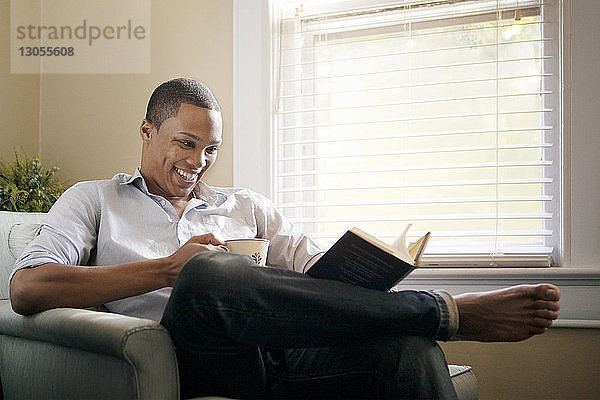 Glücklicher Mann liest Buch  während er zu Hause auf einem Sessel sitzt