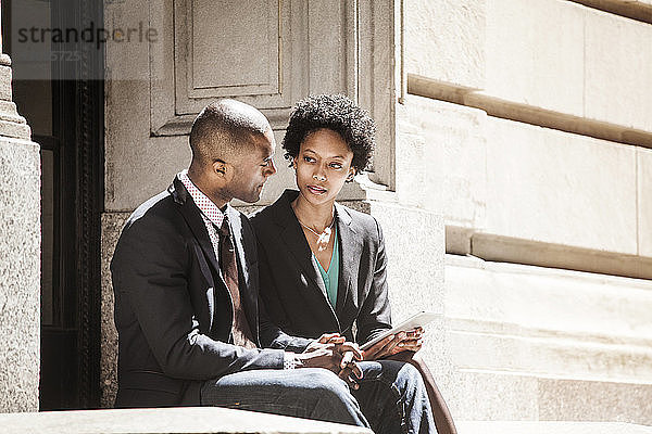 Geschäftsmann und Geschäftsfrau betrachten Tablette  während sie auf Stützmauer sitzen