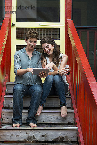 Lesbisches Paar benutzt digitales Tablett  während es auf einer Treppe vor dem Haus sitzt