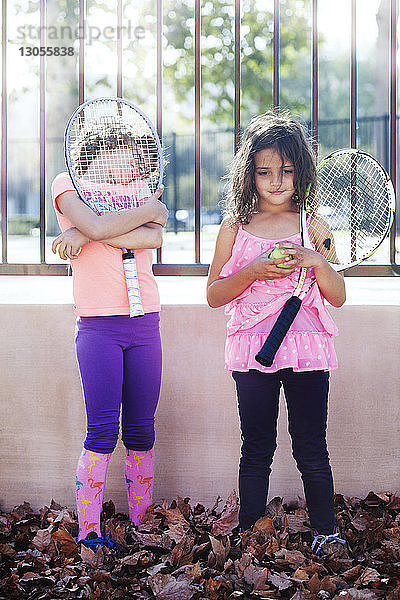 Kinder halten Tennisschläger und Ball  während sie gegen den Zaun stehen