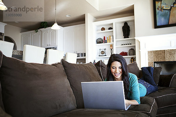 Junge Frau benutzt Laptop  während sie zu Hause auf dem Sofa liegt