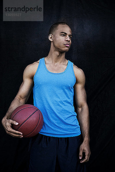 Selbstbewusster Sportler hält Basketball in der Hand  während er gegen eine schwarze Wand schaut
