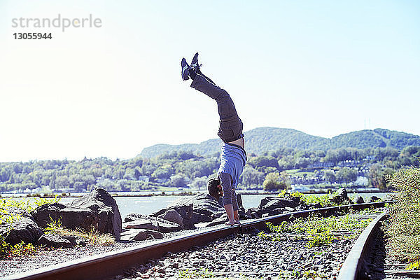 Seitenansicht eines Mannes beim Handstand auf einem Eisenbahngleis am Fluss